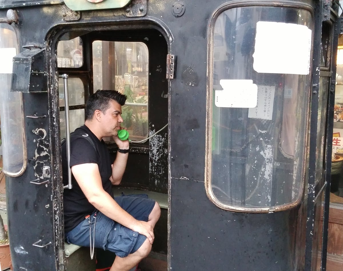 Juanjo Marcos, en cuclillas dentro de una vieja cabina telefónica negra en Japón, habla por un auricular verde chillón, con los cristales curvos de la cabina llenos de avisos escritos en japonés.