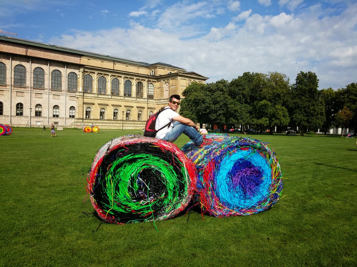 Juanjo Marcos, con gafas de sol y mochila al hombro, se sienta encima de dos enormes balas cilíndricas hechas de tiras de plástico de colores, en el césped de un parque con un gran edificio neoclásico al fondo.