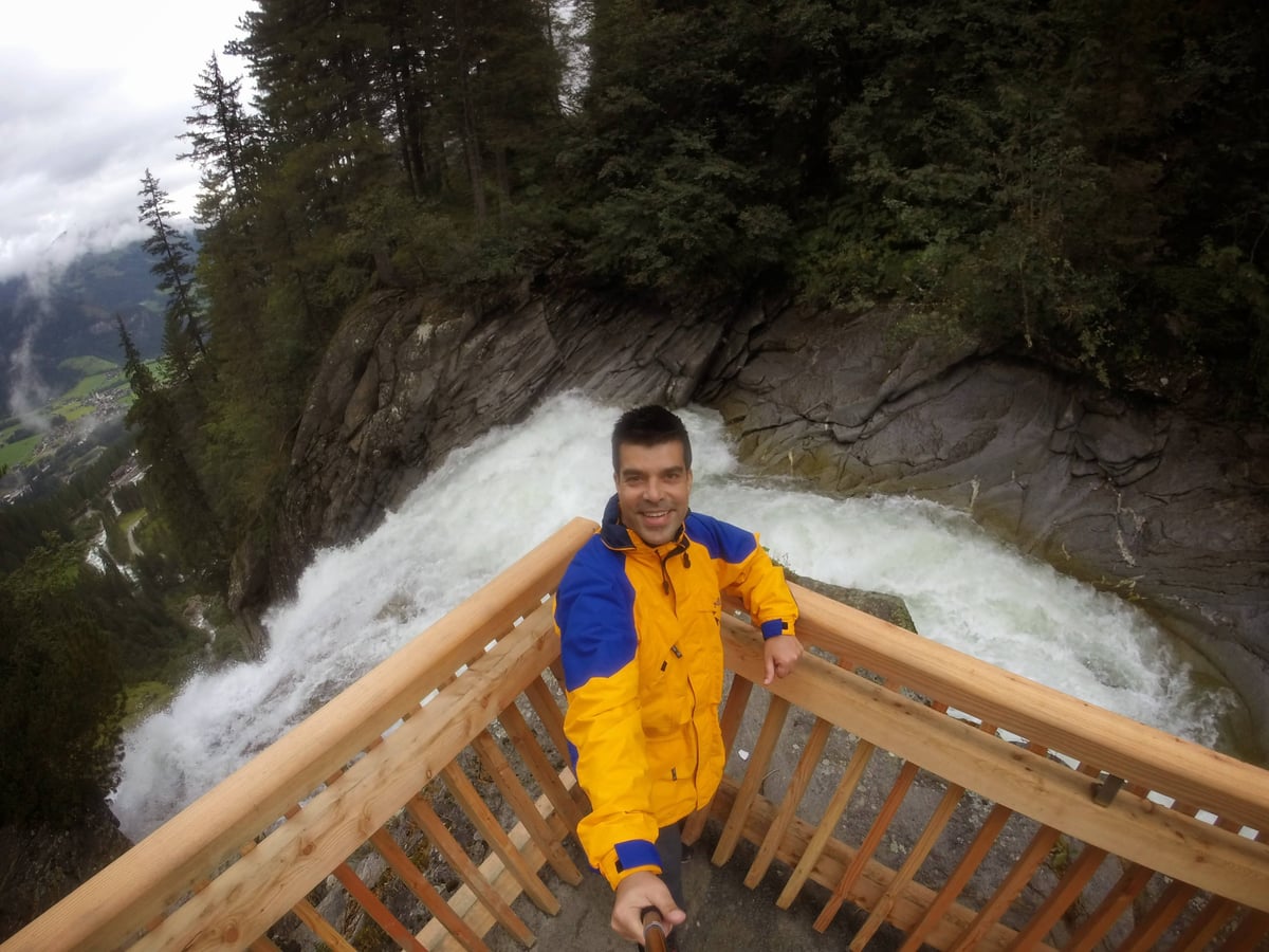 Juanjo Marcos se hace un selfie con palo, sonriendo con un chubasquero amarillo y azul, apoyado en la barandilla de madera de un mirador con una gran cascada alpina despeñándose justo detrás.
