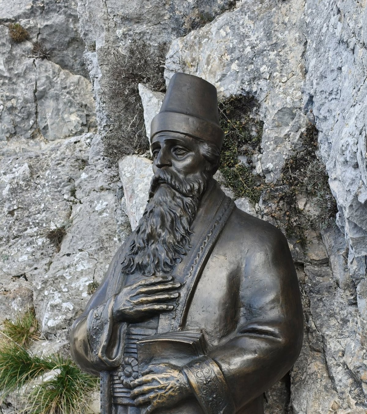 Estatua de un hombre con vestimenta tradicional junto a una pared de piedra.