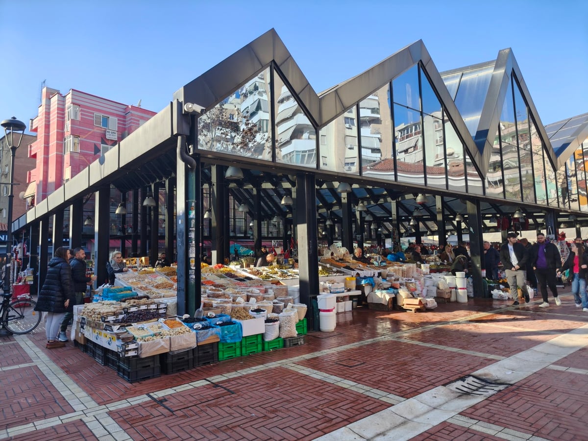 El mercado nuevo de Tirana bajo su cubierta de vidrio en diente de sierra, con los puestos de frutos secos, aceitunas y legumbres a granel en primer término y los bloques de viviendas reflejados en el techo.