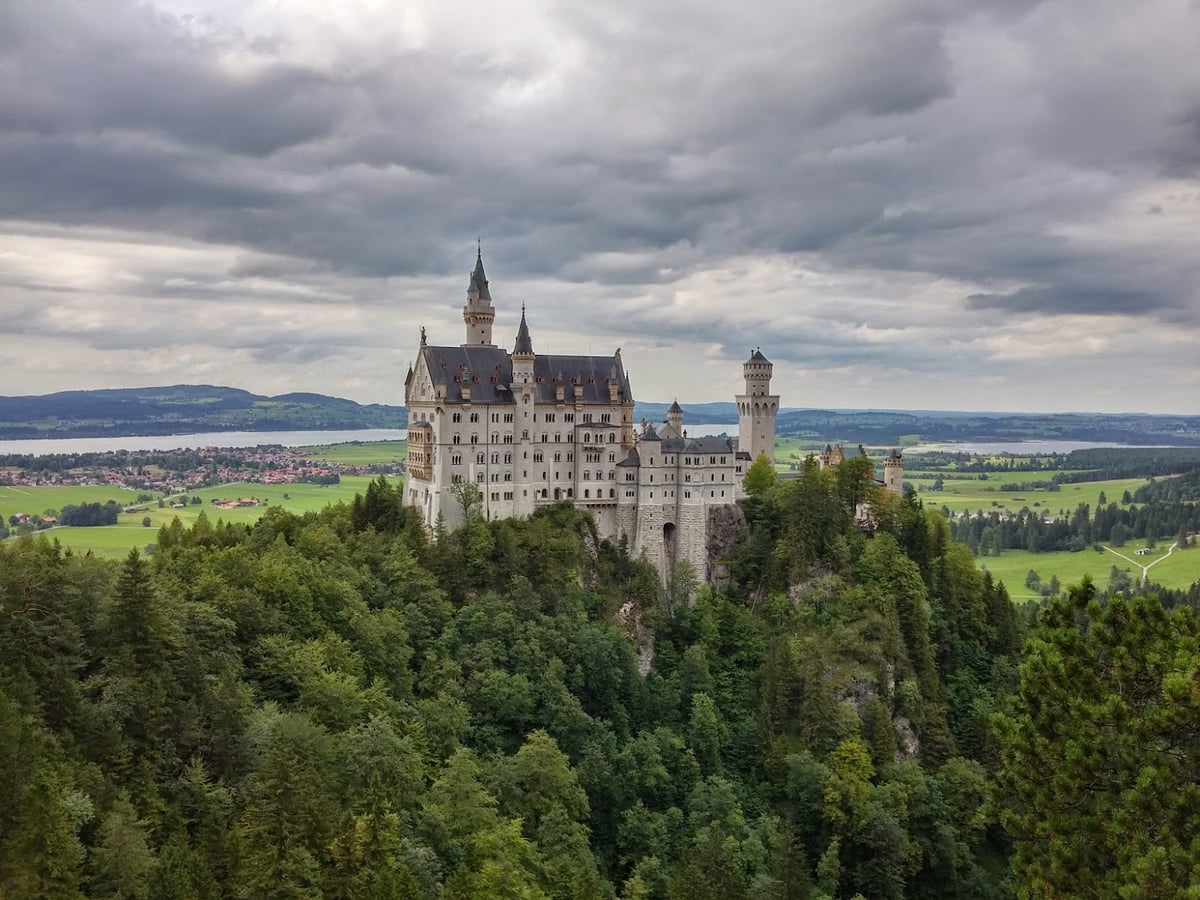 El castillo de Neuschwanstein, blanco y erizado de torres y pináculos, encaramado sobre un promontorio boscoso, con el valle verde, un lago y las colinas de Baviera extendiéndose bajo un cielo de nubes oscuras.