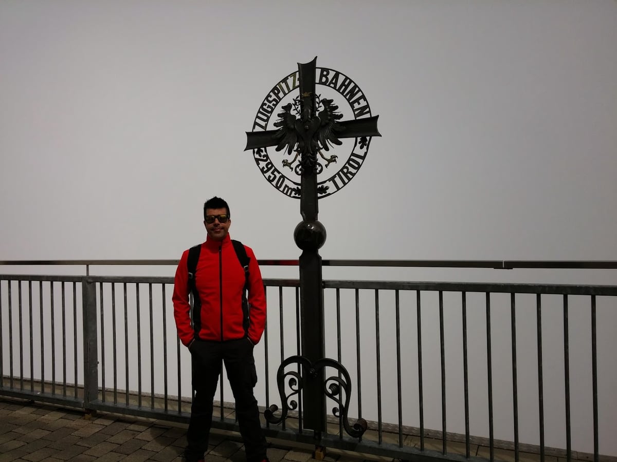Persona junto a una barandilla con una cruz decorativa y vistas a un paisaje montañoso cubierto de niebla.