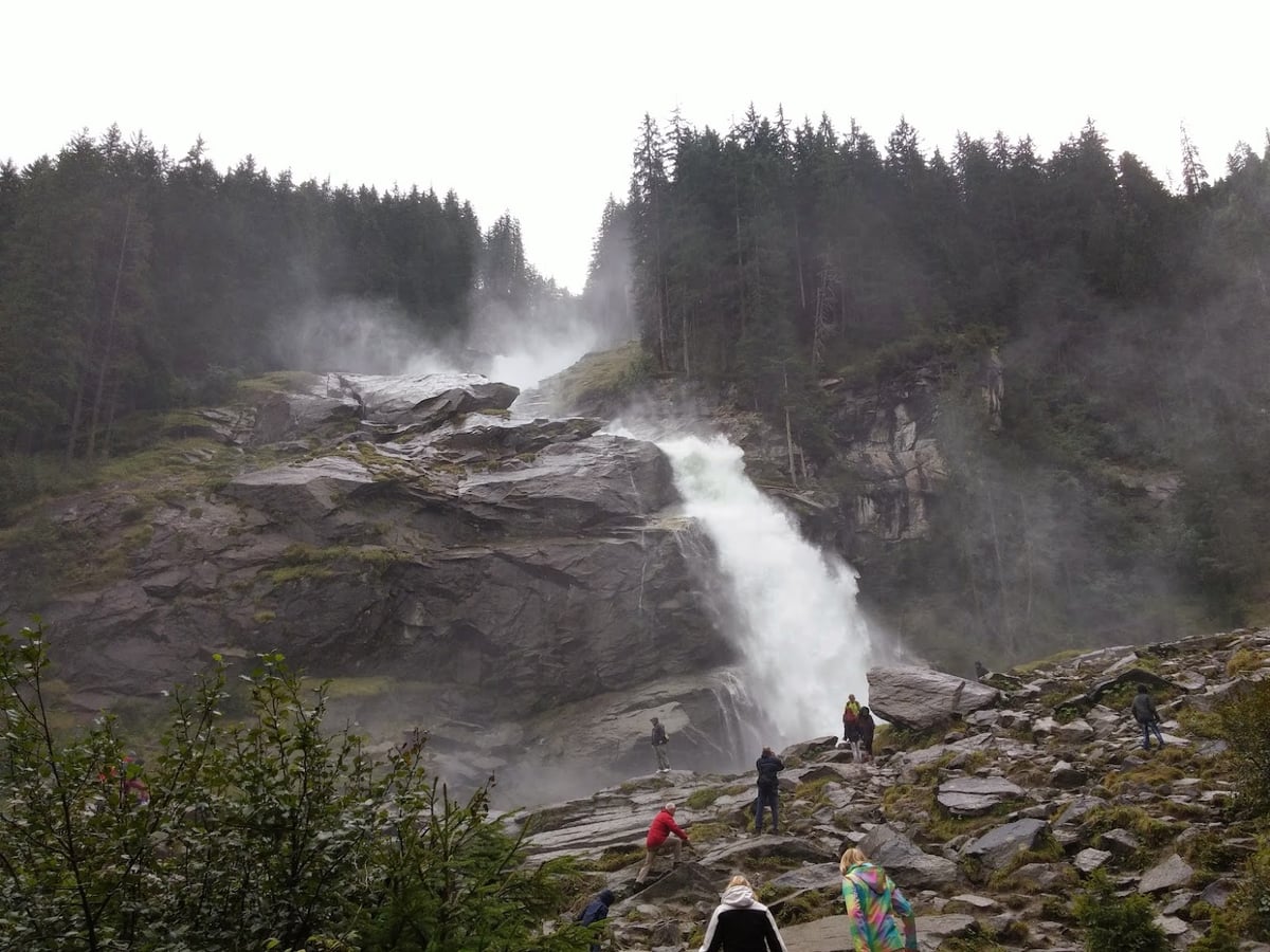 La cascada de Bad Gastein cayendo entre rocas y bosque de coníferas, con visitantes observando desde las rocas entre la niebla del agua.