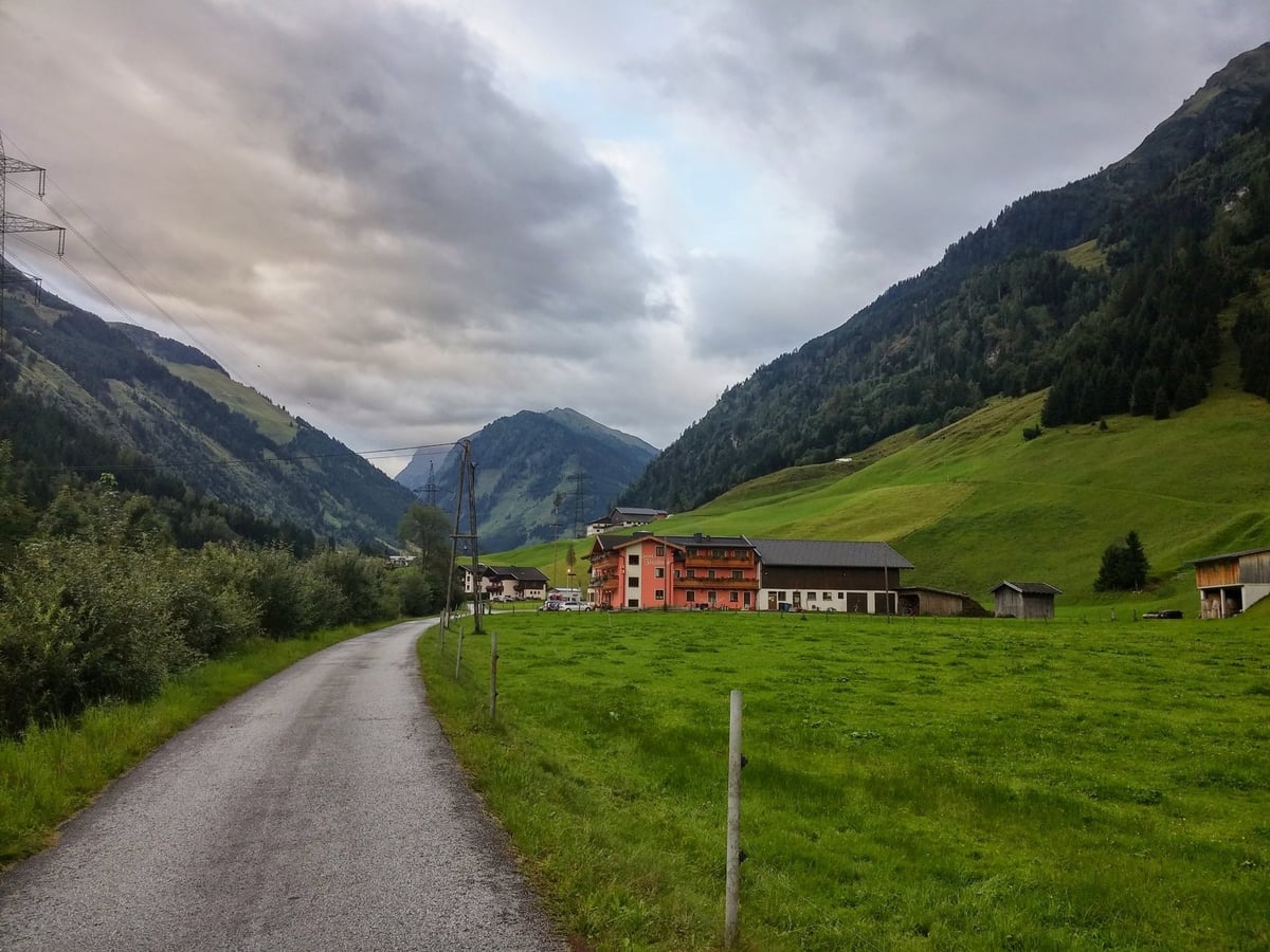 Una carretera rural serpenteando por un valle alpino, con una casa de fachada naranja y montañas verdes a ambos lados.