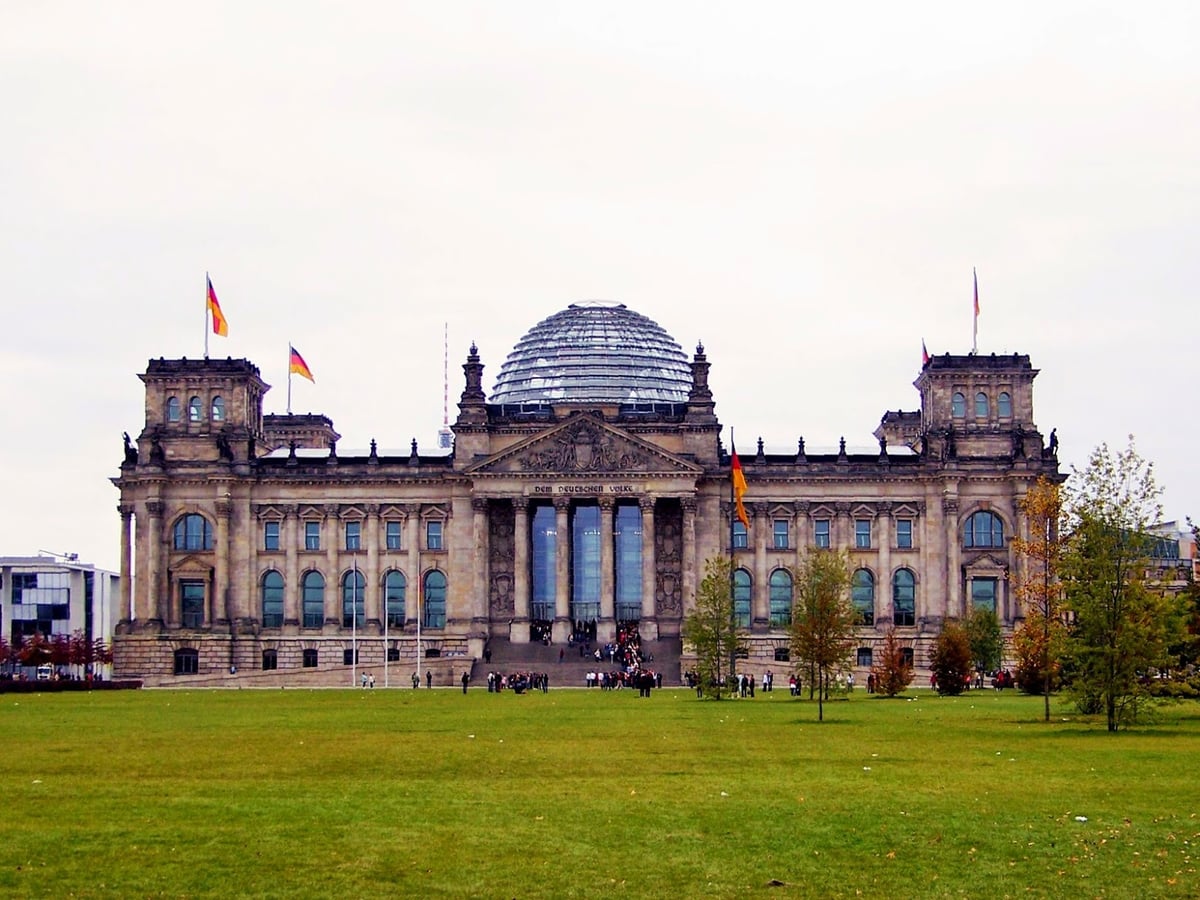 La fachada del Reichstag de Berlín vista desde el césped, con las columnas y el frontón de piedra bajo la inscripción «Dem Deutschen Volke», la cúpula de cristal asomando por detrás y cuatro banderas alemanas ondeando.