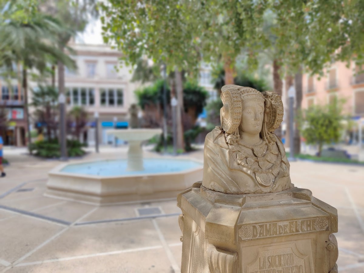 Réplica en piedra del busto de la Dama de Elche sobre un pedestal, muy enfocada en primer plano, con una plaza arbolada y su fuente circular desenfocadas al fondo.