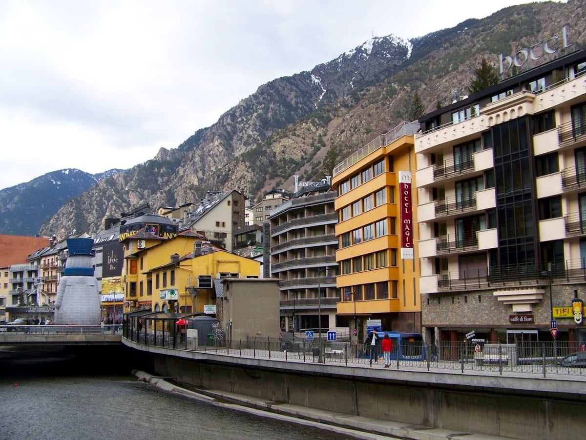 Los edificios de Andorra la Vella alineados junto al cauce encajonado del río, con hoteles y bloques modernos apretados unos contra otros y la ladera rocosa de la montaña, con algo de nieve arriba, cerrando el fondo.