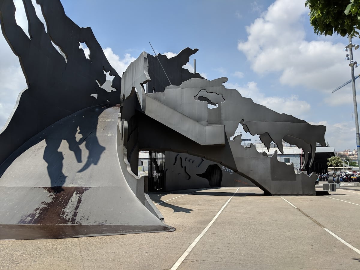 Enorme escultura de acero oscuro recortado con formas orgánicas, que se pliega sobre sí misma formando un pórtico sobre una explanada de hormigón, con su sombra dibujada en el suelo.
