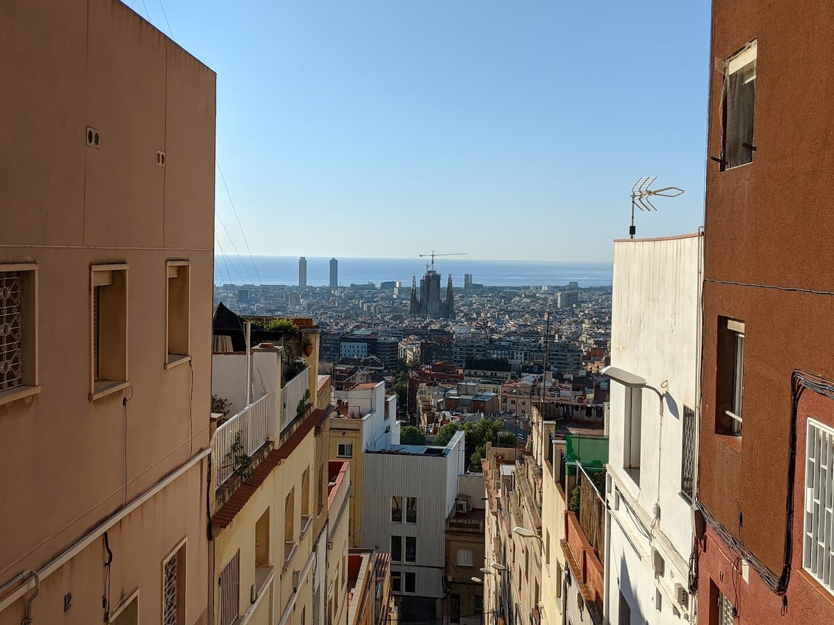 Vista de Barcelona encajada entre las medianeras de dos edificios de una calle en cuesta, con la Sagrada Familia y sus grúas en el centro, los tejados extendiéndose hasta el puerto y el mar cerrando el horizonte.