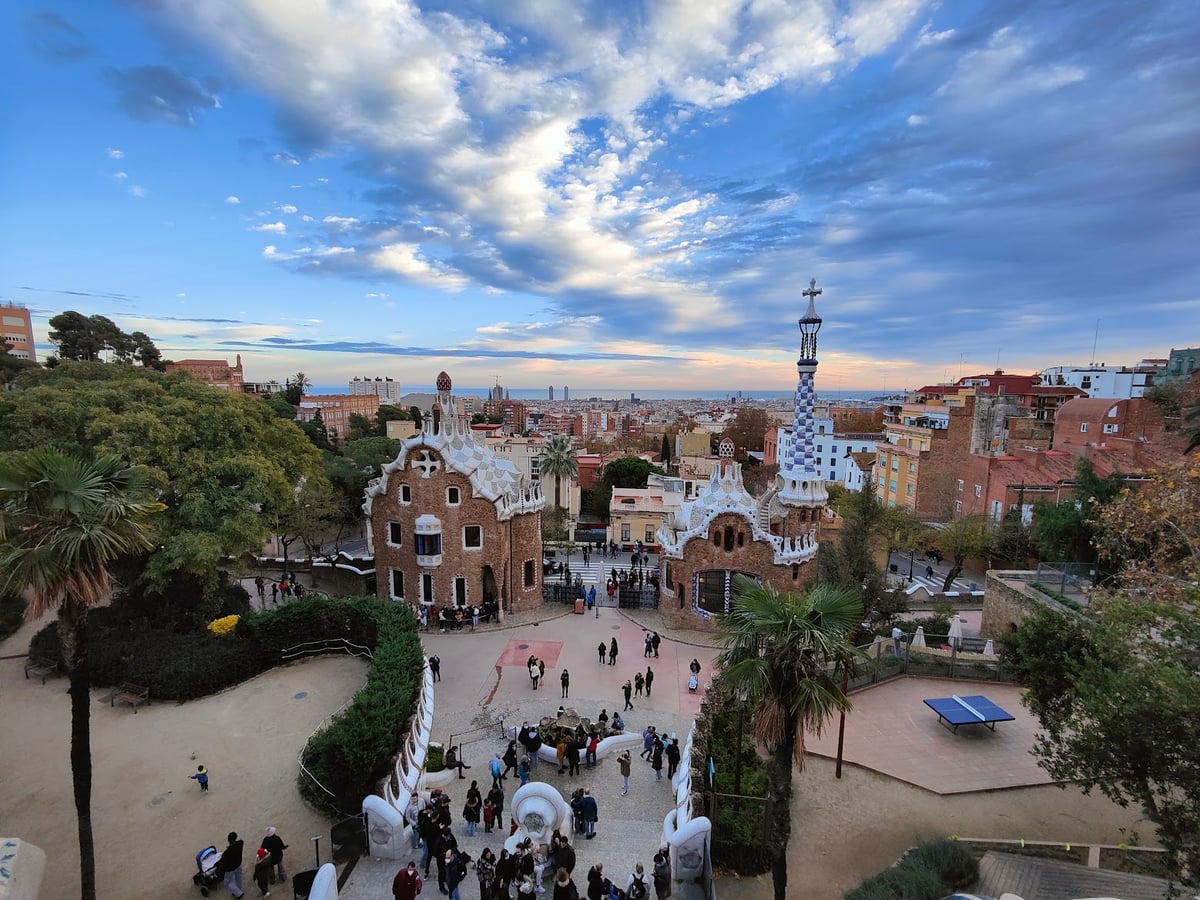 El Park Güell desde lo alto, con los dos pabellones de Gaudí de tejados ondulados y cerámica blanca flanqueando la entrada, la gente subiendo por la escalinata y Barcelona y el mar al fondo bajo un cielo de nubes.