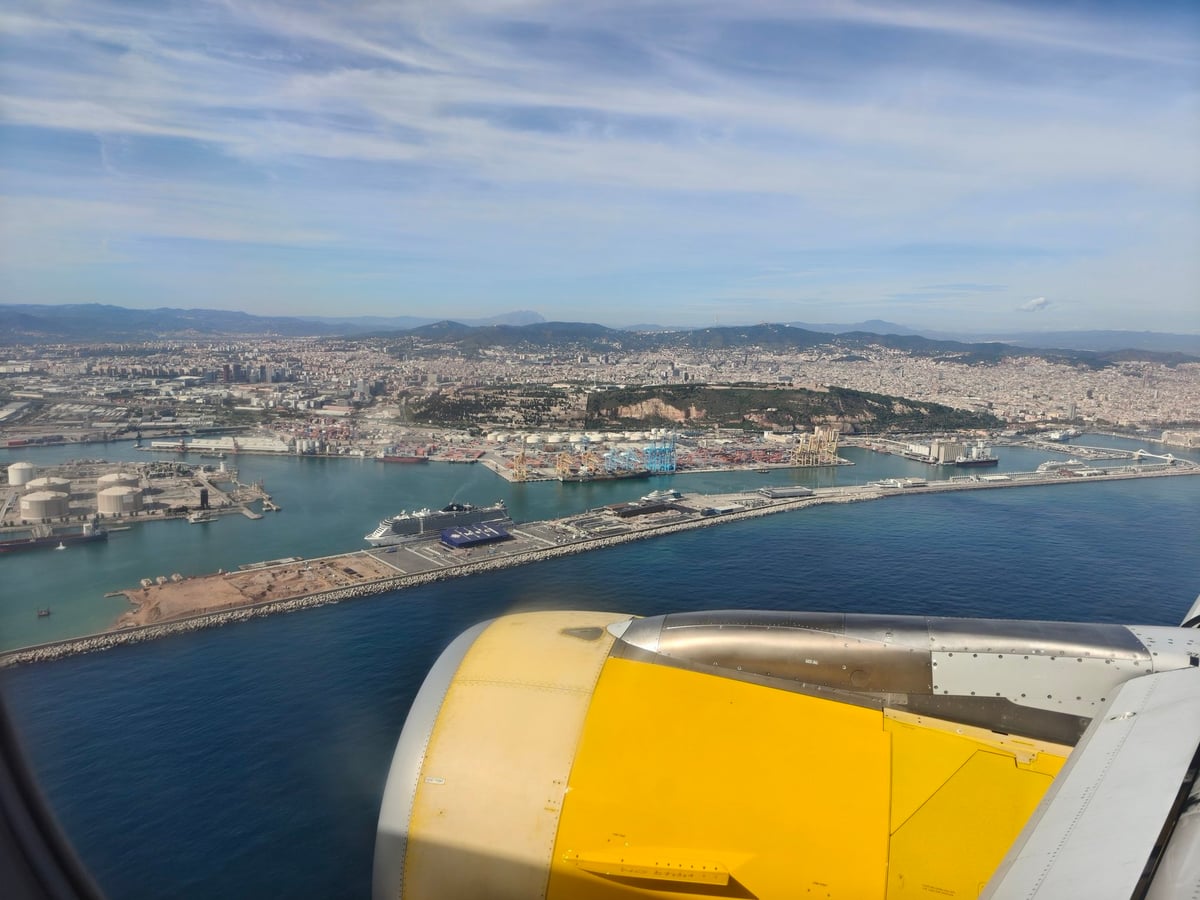 El puerto de Barcelona visto desde la ventanilla del avión, con barcos y grúas, la ciudad al fondo y el motor amarillo del avión en primer plano.