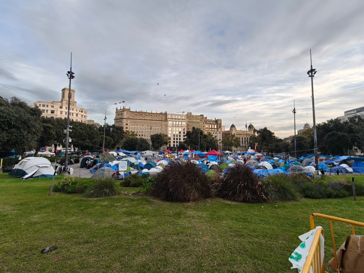 Tiendas de campaña de colores que llenan la plaza de Catalunya, con matas y una valla amarilla en primer plano y los edificios de la plaza al fondo bajo un cielo nublado.