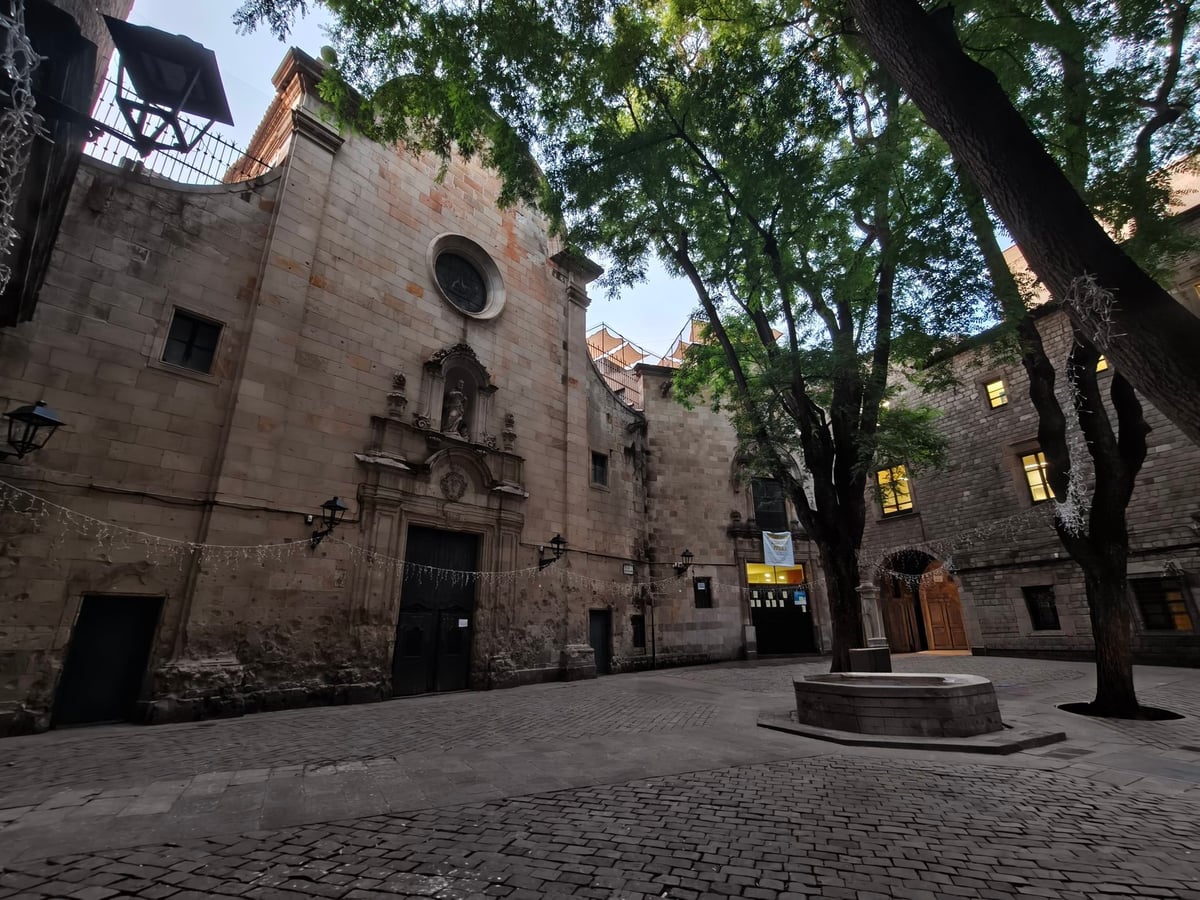 La plaza de Sant Felip Neri, con la fachada de piedra de la iglesia a la izquierda, una pequeña fuente octogonal y los árboles sobre el empedrado.