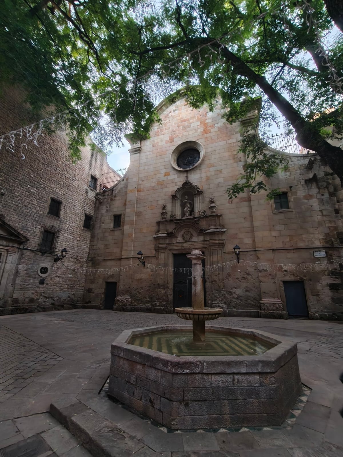 Fachada de la iglesia de Sant Felip Neri con su rosetón, vista desde la fuente octogonal de la plaza, entre las copas de los árboles.