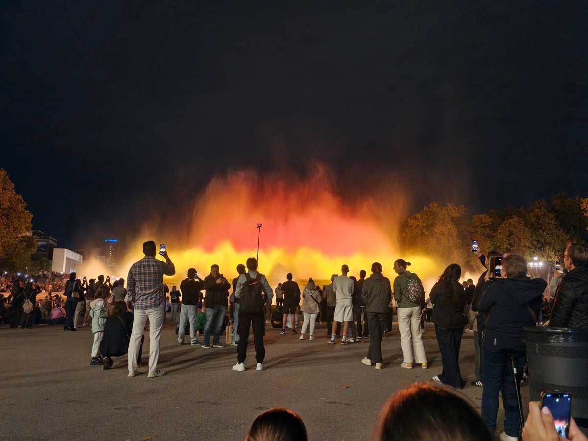 Chorros de agua iluminados de naranja y amarillo en la Font Màgica de Montjuïc, con decenas de personas de pie mirando y fotografiando el espectáculo.