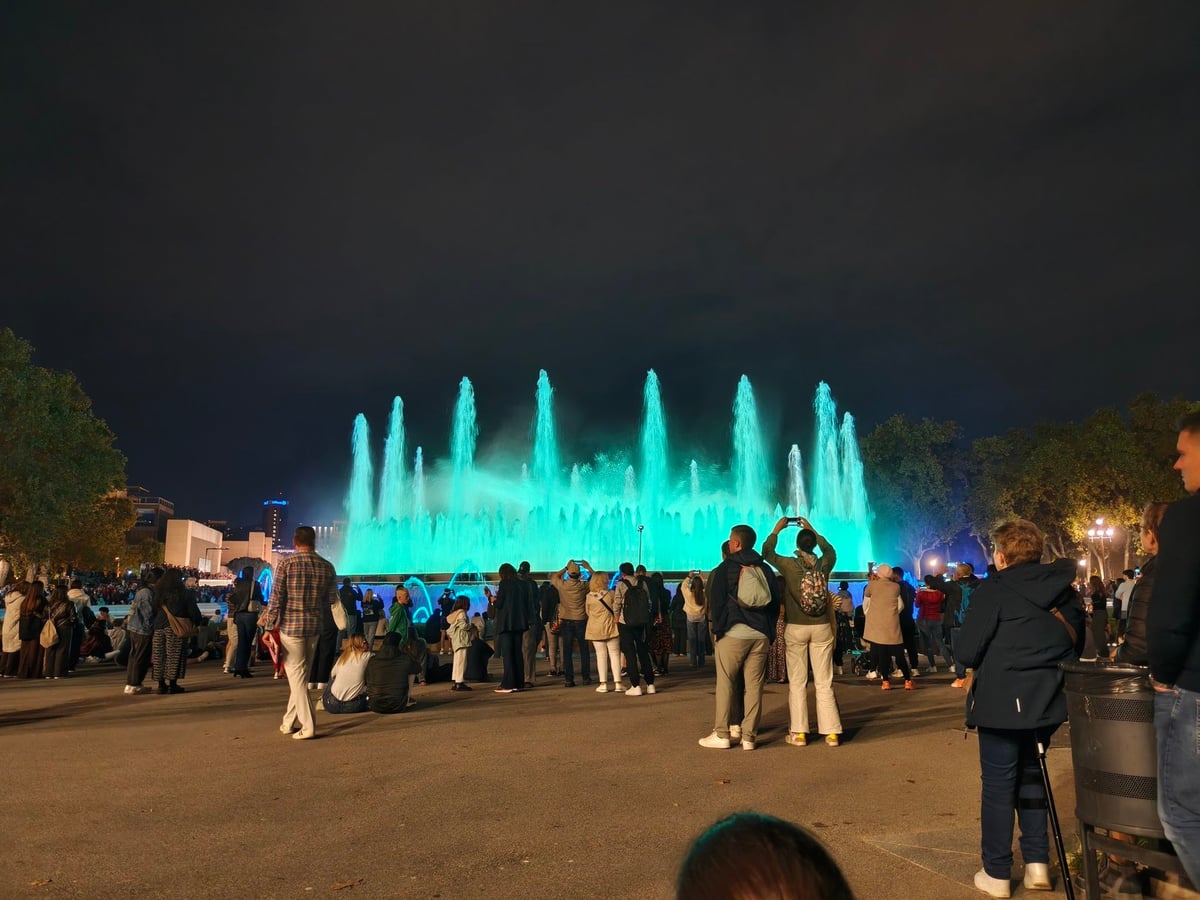 Los chorros de la Font Màgica iluminados de turquesa en la noche, con espectadores de pie alrededor.