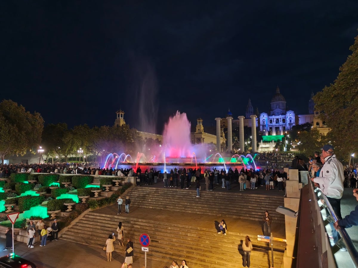 La fuente con chorros rosas y verdes vista desde lo alto de las escaleras, con la multitud en las gradas y el Palau Nacional iluminado al fondo.