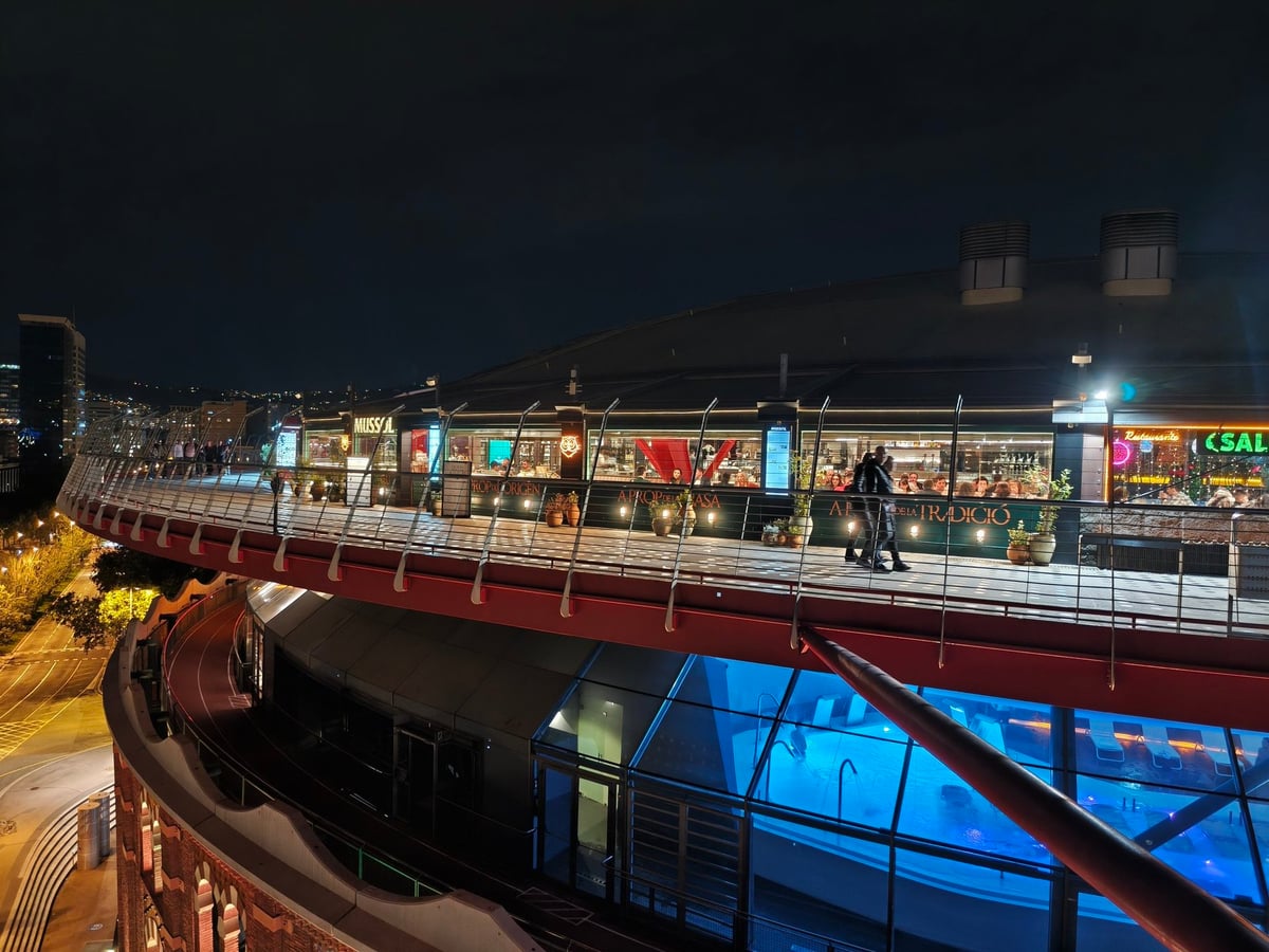 Anillo exterior de la terraza de Las Arenas de noche, con restaurantes iluminados en la planta superior y una gran cúpula de cristal azulada debajo.