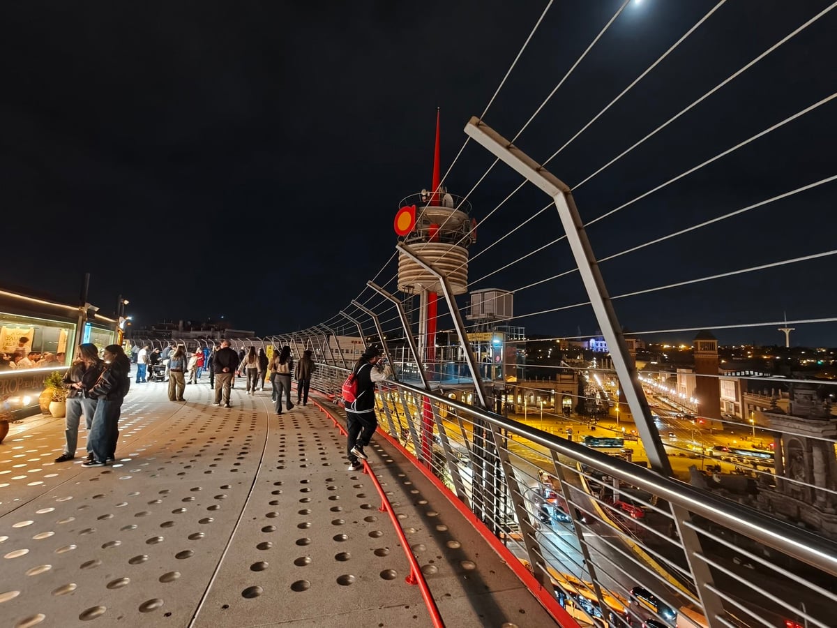 Pasarela de la terraza de Las Arenas con suelo perforado, barandilla de cables de acero y la ciudad iluminada al fondo.