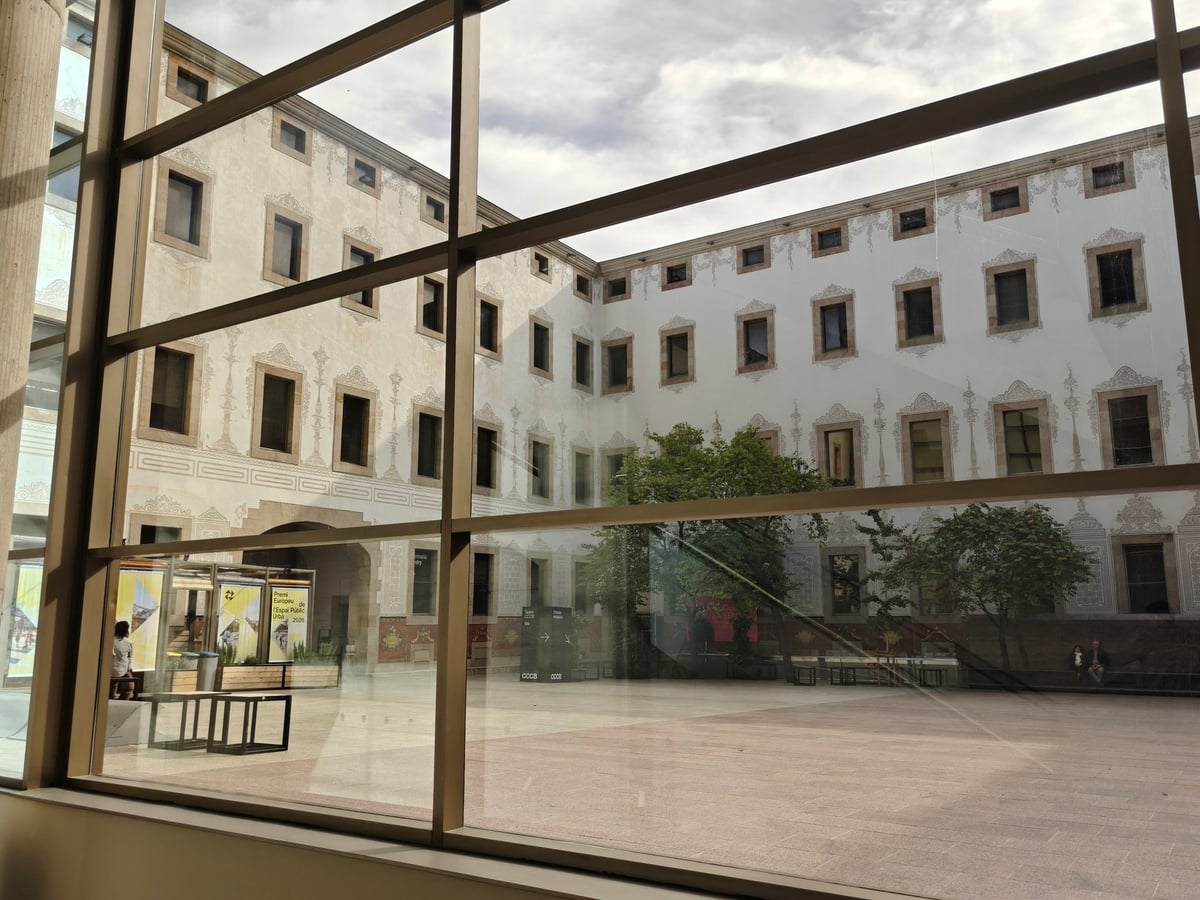 Patio del edificio antiguo del CCCB, con sus fachadas blancas de ventanas sencillas, visto desde el edificio moderno a través de una cristalera con reflejos.