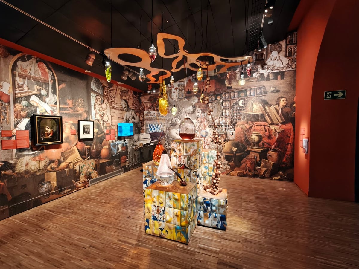 Sala de la exposición con las paredes cubiertas por grabados antiguos de gabinetes y laboratorios, objetos de vidrio y azulejos sobre plintos y una pieza naranja calada colgada del techo.
