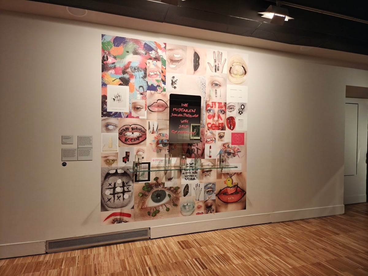 Mural en collage con ojos, labios, manos y recortes de publicidad de cosmética sobre una pared blanca de la exposición, con un cartel negro de texto en el centro.