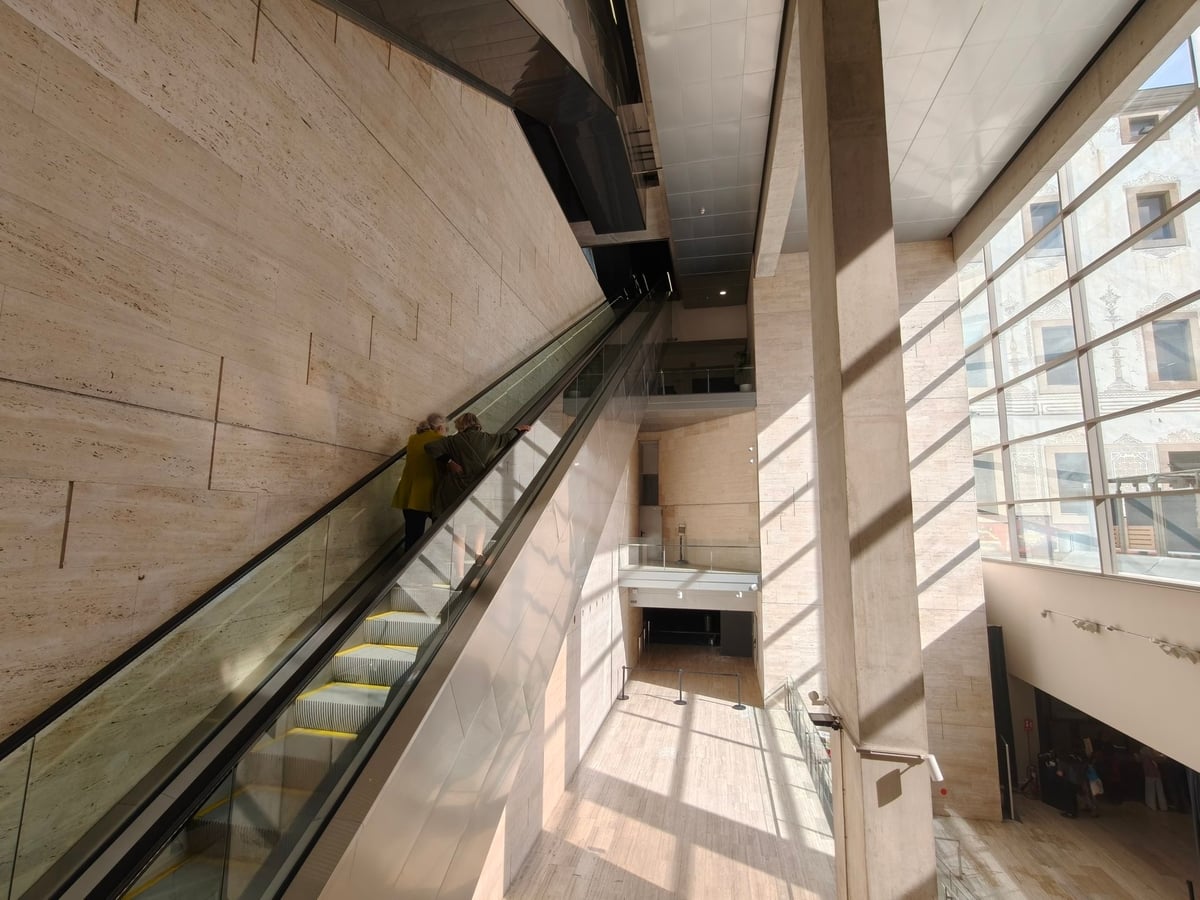 Escalera mecánica de cristal que sube junto a una pared de travertino, con dos personas mayores de espaldas en los escalones y una gran cristalera a la derecha por la que entra el sol y se ven las fachadas esgrafiadas de enfrente.