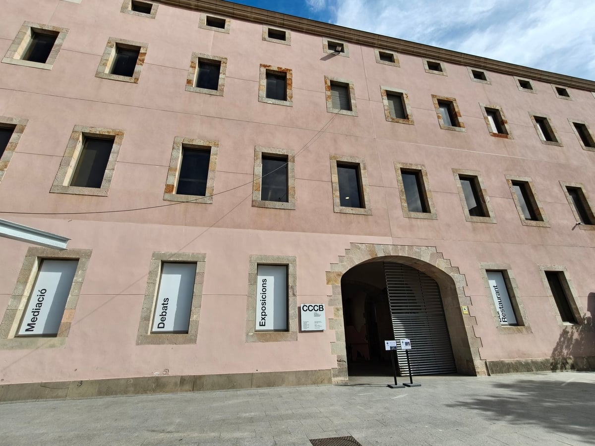 Fachada rosa de la antigua Casa de la Caritat, con ventanas enmarcadas en piedra, un arco de sillería con persiana metálica como entrada y carteles blancos en las ventanas bajas que dicen «Mediació», «Debats», «Exposicions» y «Restaurant», junto a la placa del CCCB.