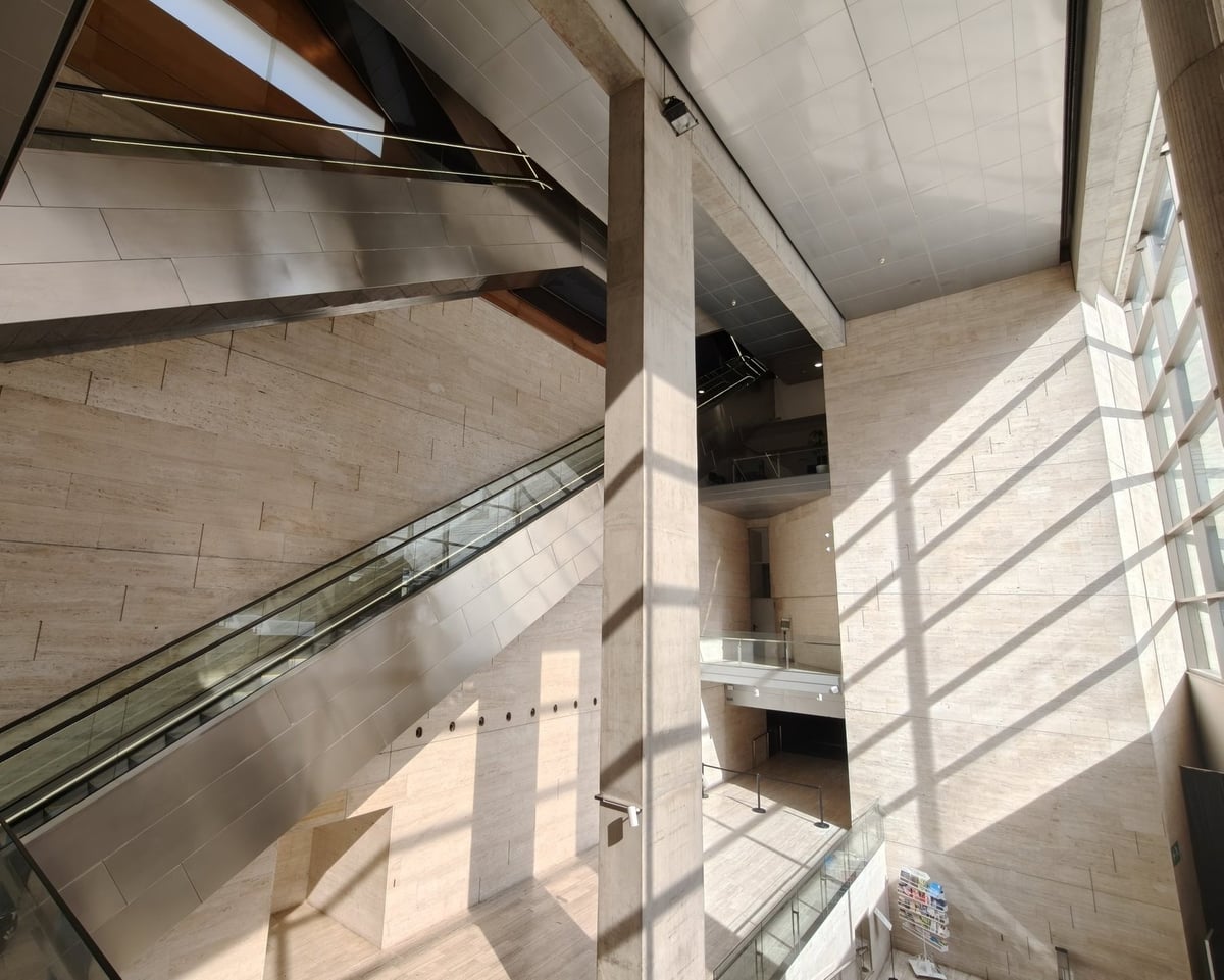 Vista desde lo alto del vestíbulo del CCCB: la escalera mecánica forrada de acero cruza el espacio, con un pilar de hormigón en el centro y las sombras de la cristalera dibujadas sobre las paredes de travertino.