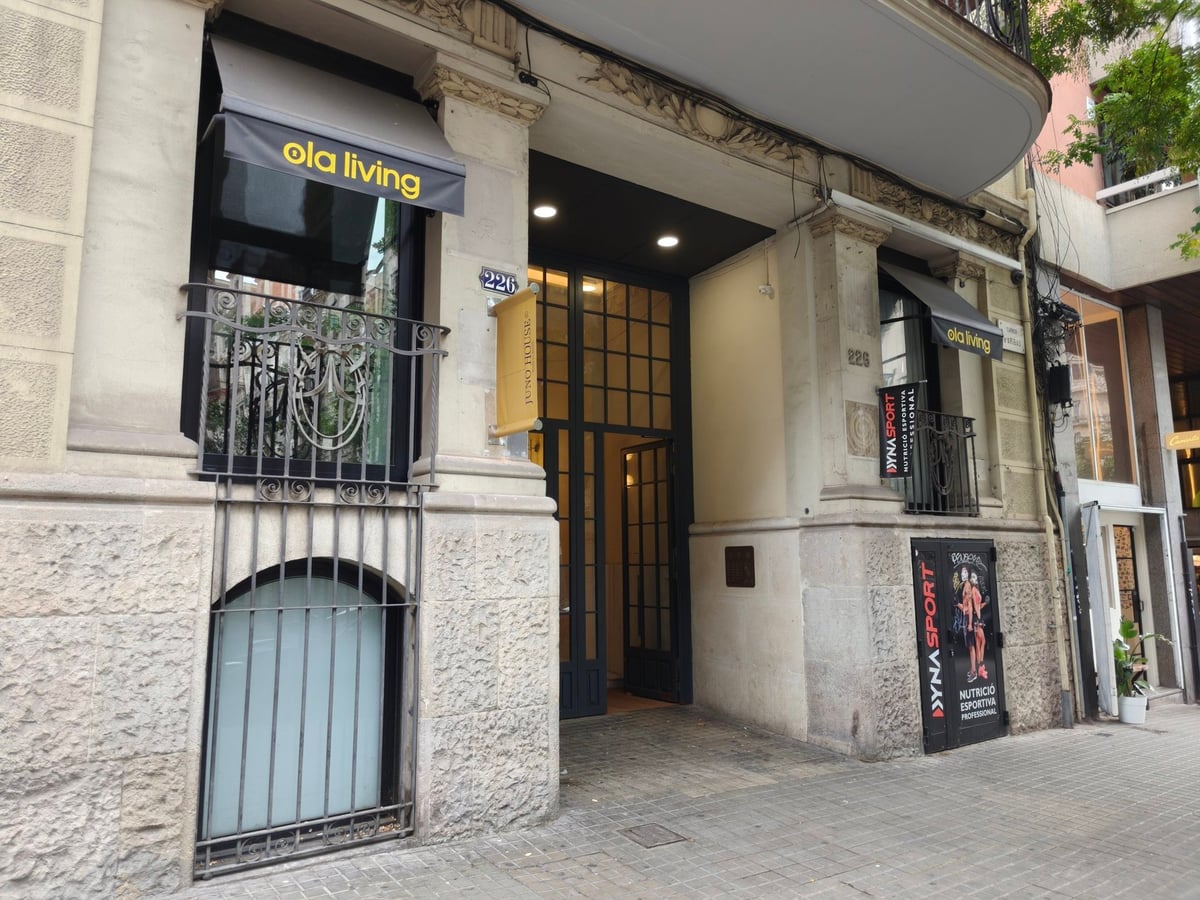 Portal de piedra con puerta de hierro y cristal y el rótulo «ola living» en un toldo, en una calle de Barcelona.