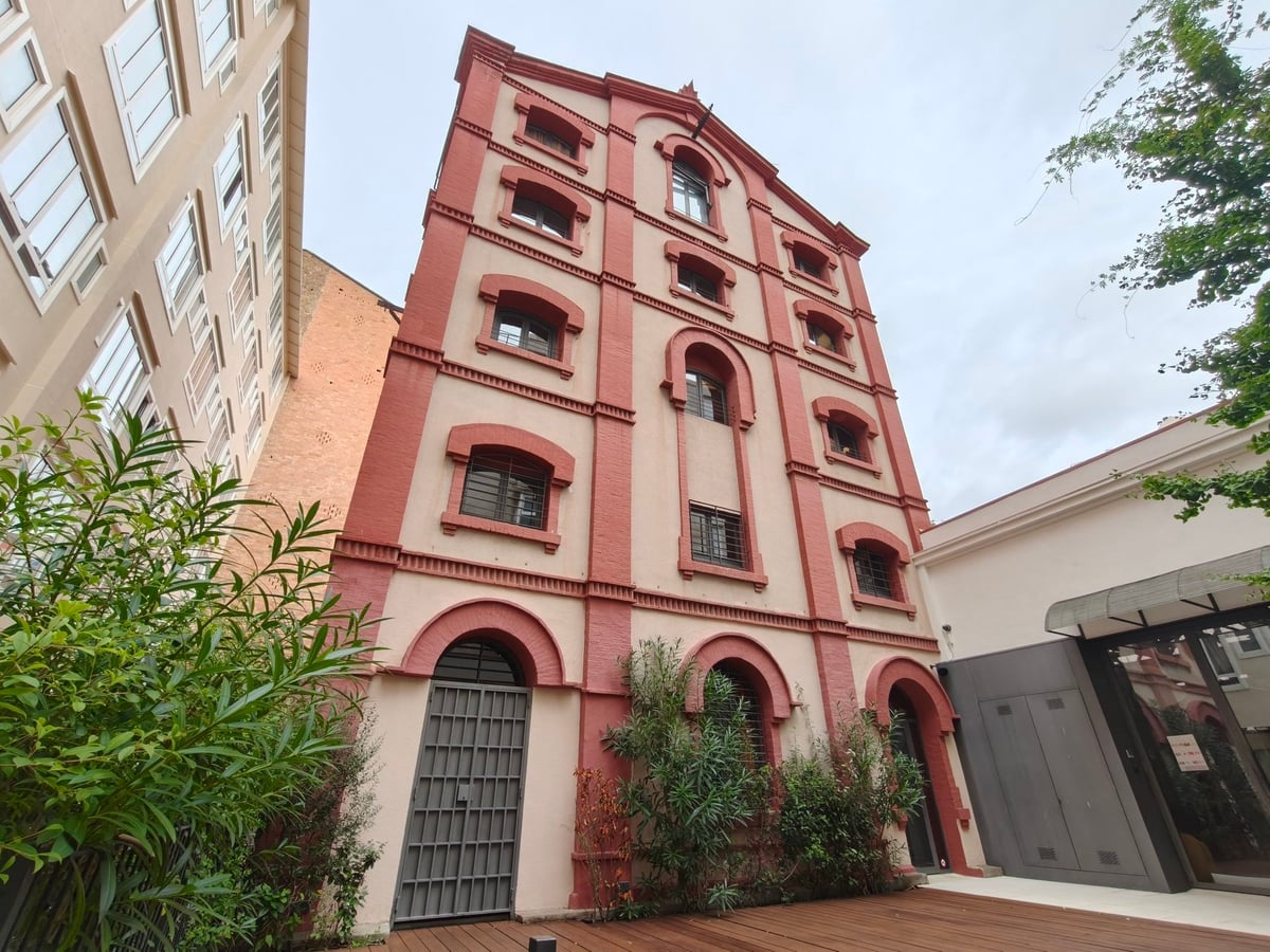 Edificio alto y estrecho de fachada rojiza con ventanas de arco, en un patio interior con vegetación y suelo de madera.