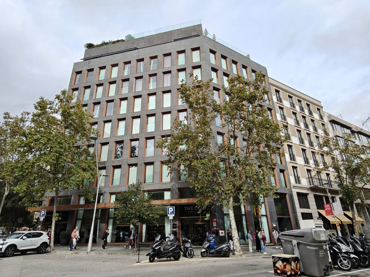 Fachada del hotel The One Barcelona, un edificio de cristal y piedra gris con hileras de ventanas, en una esquina rodeada de árboles.