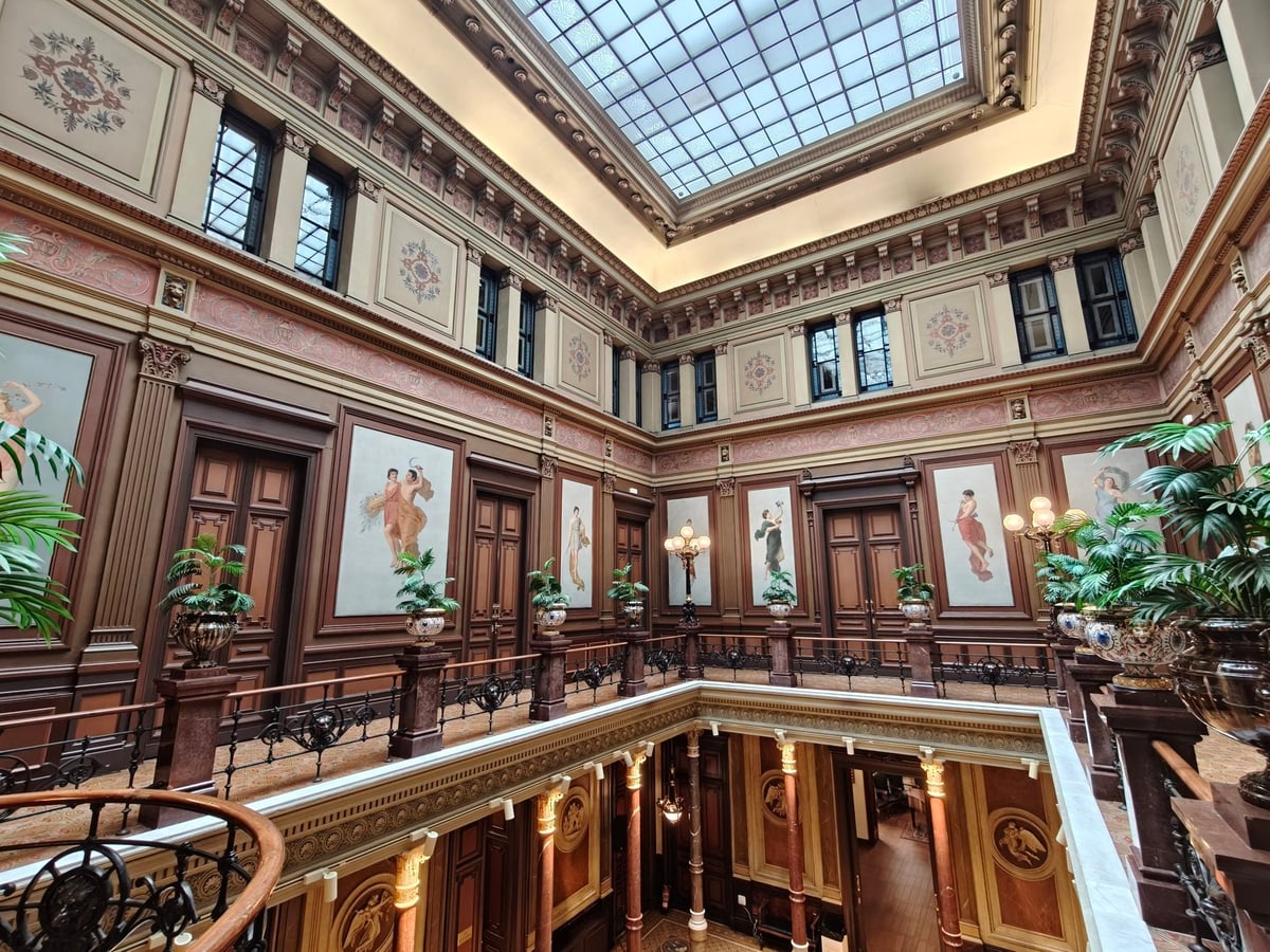 Galería superior del mismo patio, con paneles pintados con figuras entre puertas de madera, plantas y la claraboya iluminando la sala.