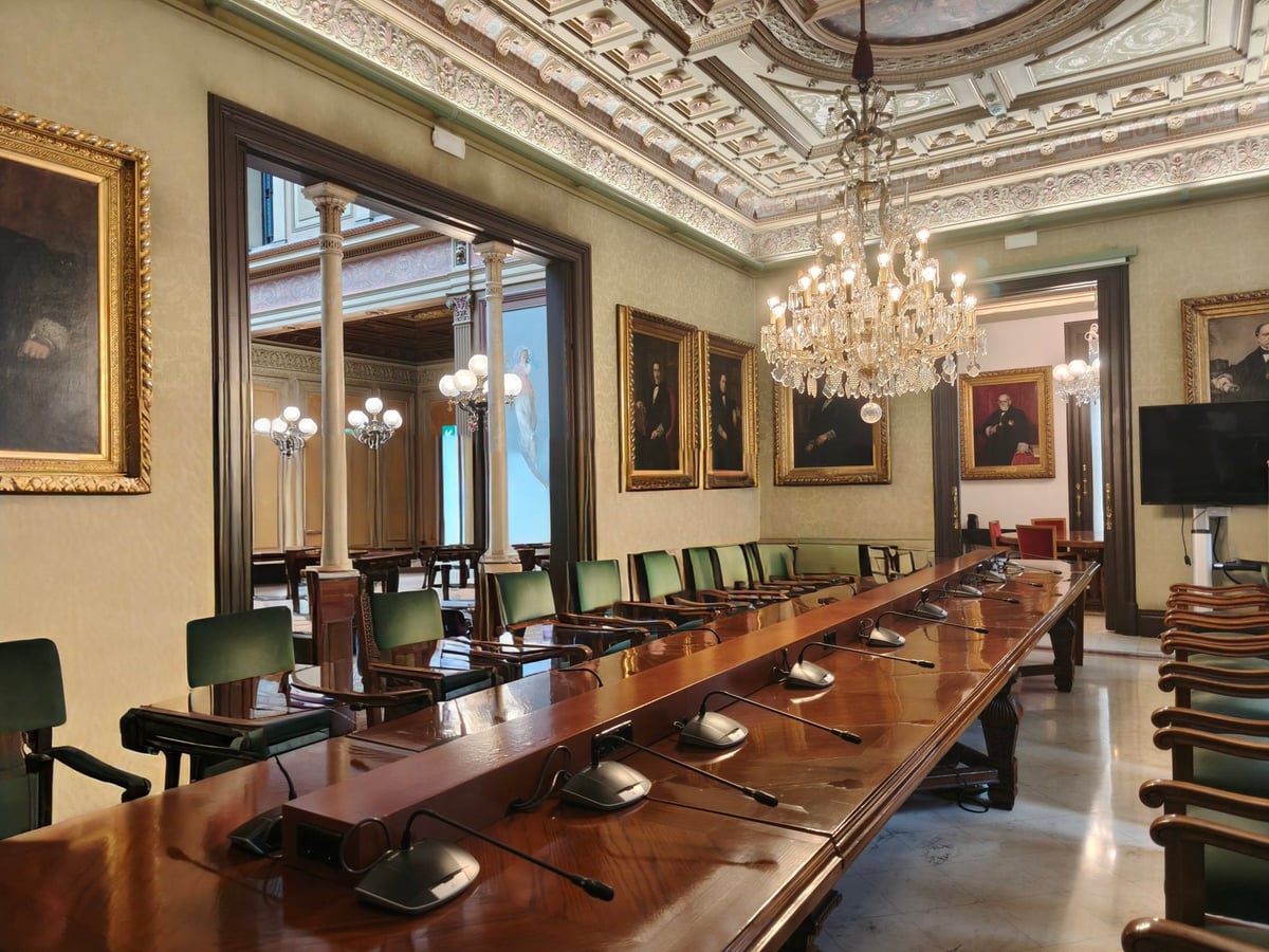 Sala con una larga mesa de madera con micrófonos y sillas verdes, retratos al óleo en marcos dorados y una lámpara de araña bajo un techo decorado.