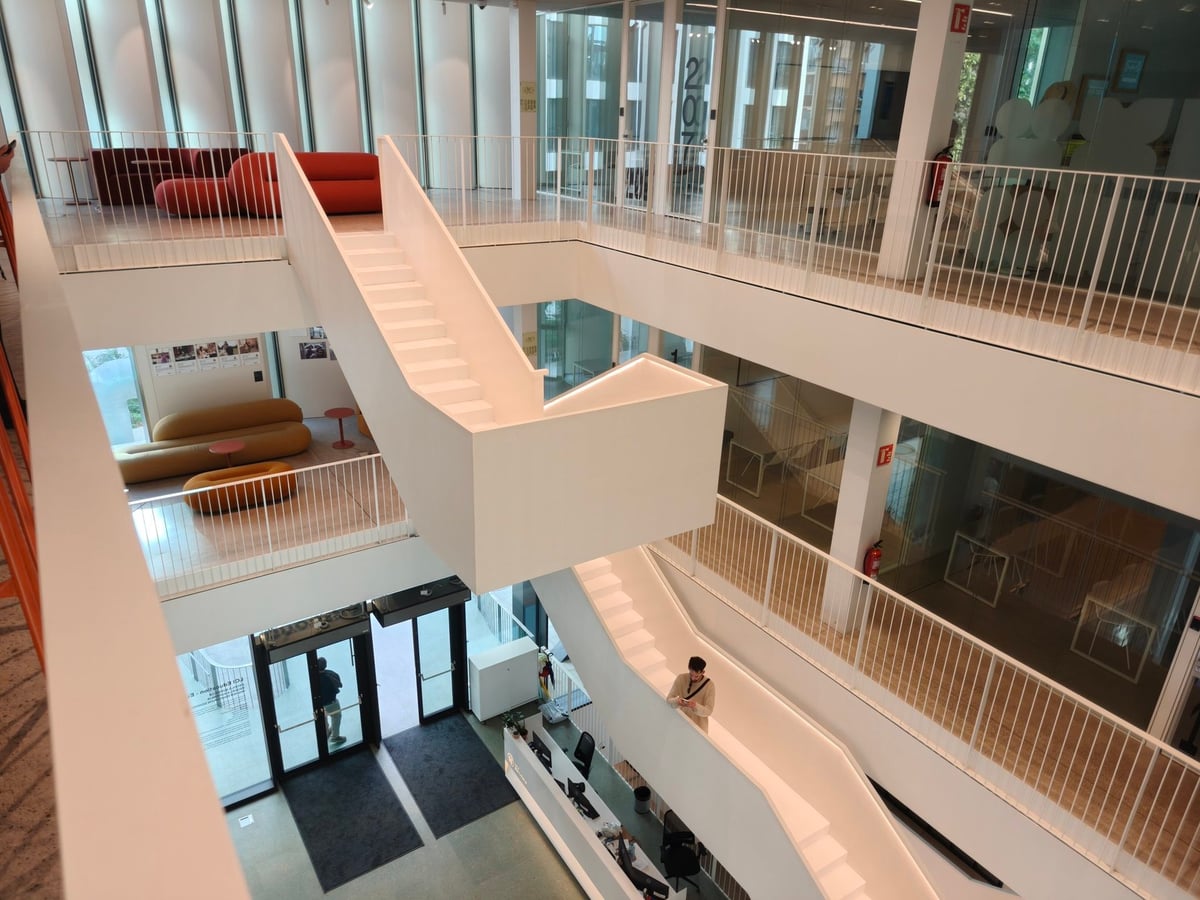 Atrio del campus con escaleras blancas que se cruzan, balcones con barandillas blancas y sofás rojos en las plantas altas.