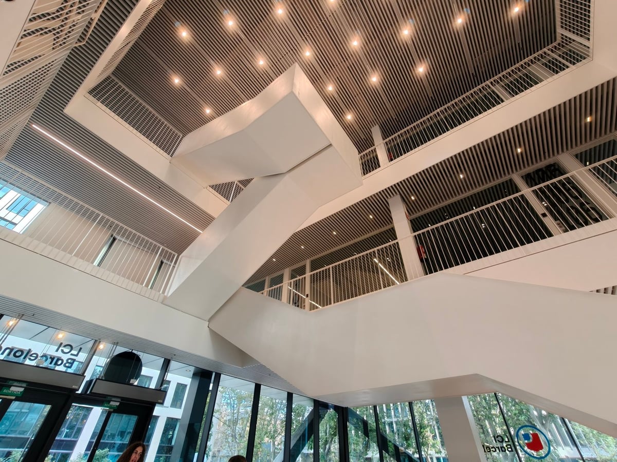 Escaleras blancas del atrio vistas desde abajo, bajo un techo de lamas con luces puntuales y con el rótulo «LCI Barcelona» en el cristal.