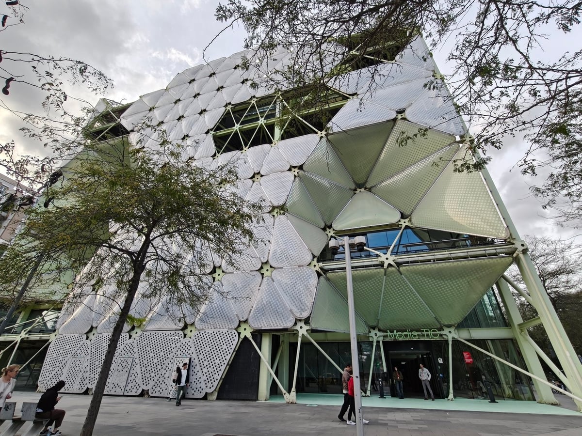Esquina del edificio Media-TIC con la fachada de almohadillas neumáticas blancas y verdes sujetas a una estructura de acero, vista desde abajo entre las ramas de un árbol.