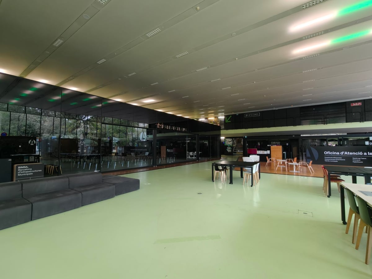 Vestíbulo del Media-TIC con suelo verde, mesas de trabajo, sofás negros y una gran cristalera, con lámparas fluorescentes alargadas en el techo.