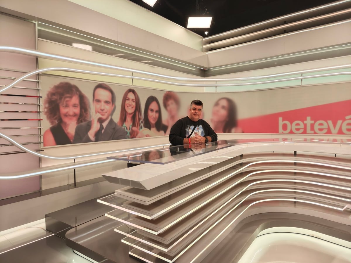 Plató de informativos de Betevé con una mesa curva de luces blancas y, al fondo, un panel con las fotografías de sus presentadores y el rótulo «betevé»; un visitante posa detrás de la mesa.