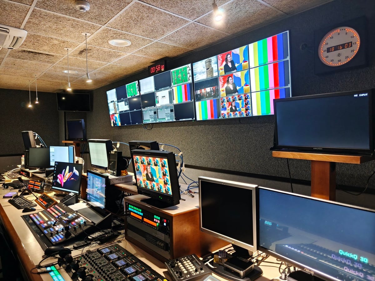 Sala de control de Betevé con una pared de monitores, mesas de mezclas y pantallas, y un reloj naranja en la pared.