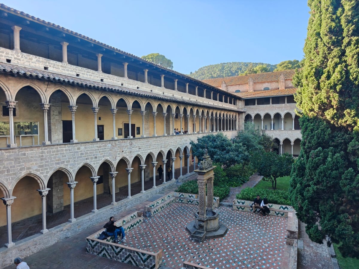 Claustro del monasterio de Pedralbes, con tres pisos de galerías con arcos, un jardín con cipreses y una fuente en el centro, y colinas al fondo.
