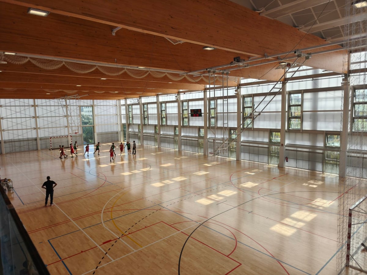 Interior del Pabellón Illa, con un techo de vigas de madera, grandes cristaleras translúcidas y jugadores en la pista al fondo.