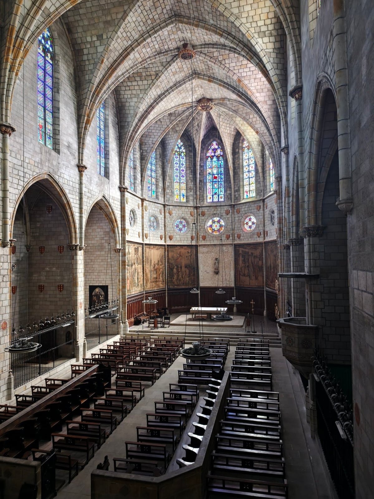 Nave gótica de la iglesia del monasterio vista desde un nivel elevado, con filas de bancos de madera, el ábside con vidrieras de colores y pinturas en los paños laterales.