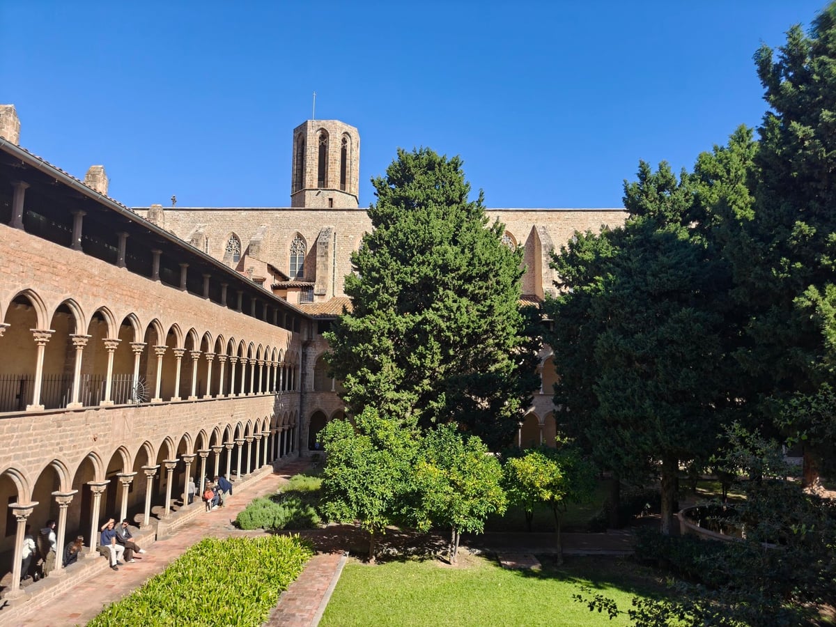 El claustro de Pedralbes con la torre del campanario al fondo, dos galerías de arcos y árboles en el jardín.