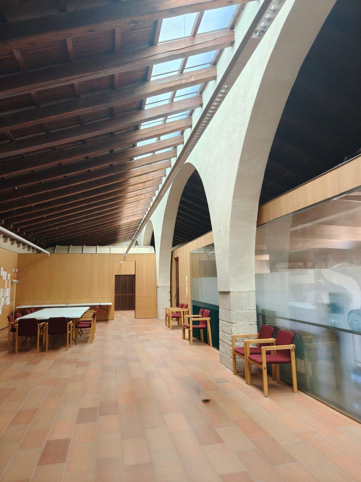 Sala del monasterio con vigas de madera vistas, una claraboya, grandes arcos de piedra, suelo de baldosa y sillas con tapizado rojo junto a una mesa.