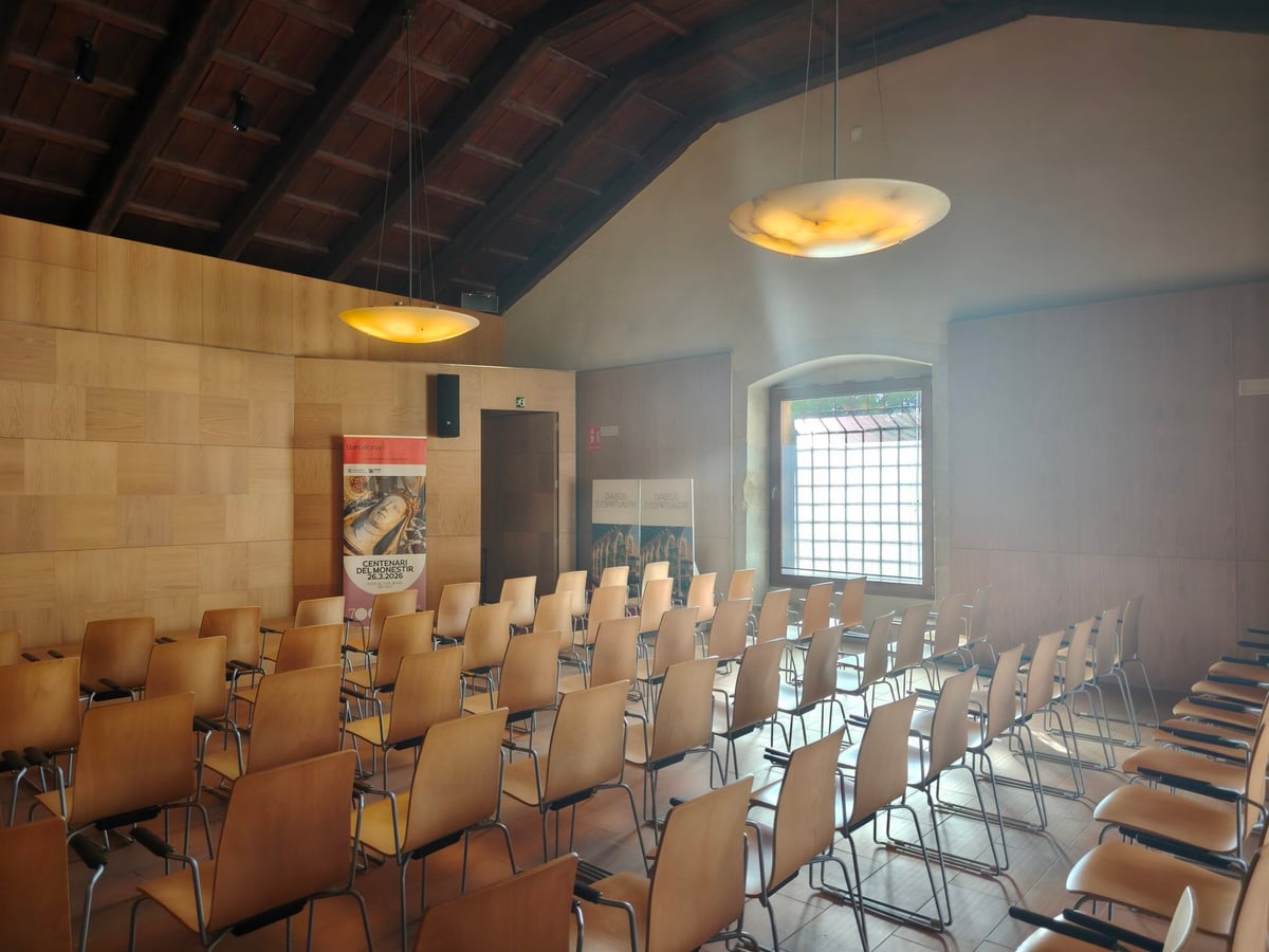 Salón con filas de sillas de madera, lámparas de pantalla colgadas del techo de vigas y un cartel enrollable a la izquierda.
