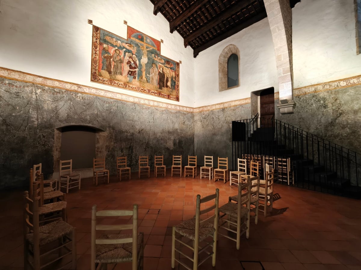 Sala del monasterio con zócalo pintado en tonos verdosos, un tapiz en la pared, sillas de enea en círculo y una escalera de hierro a la derecha.