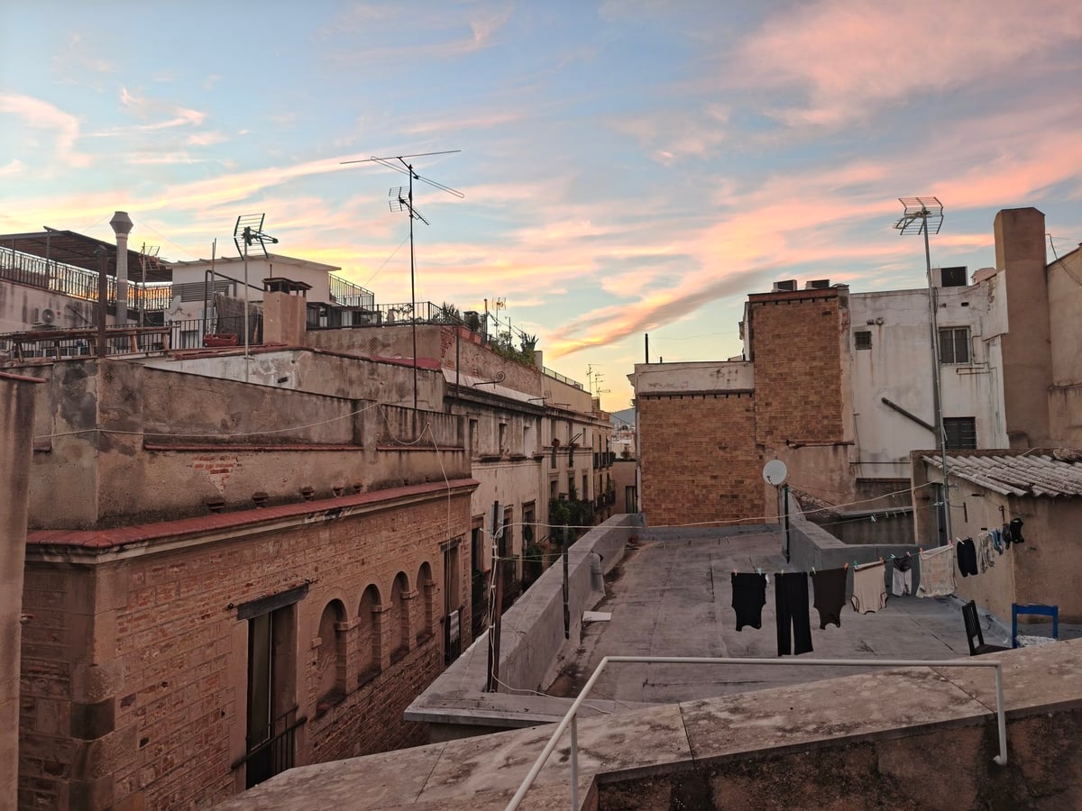 Tejados y azoteas de Barcelona al atardecer, con antenas, ropa tendida en una terraza y nubes rosadas.
