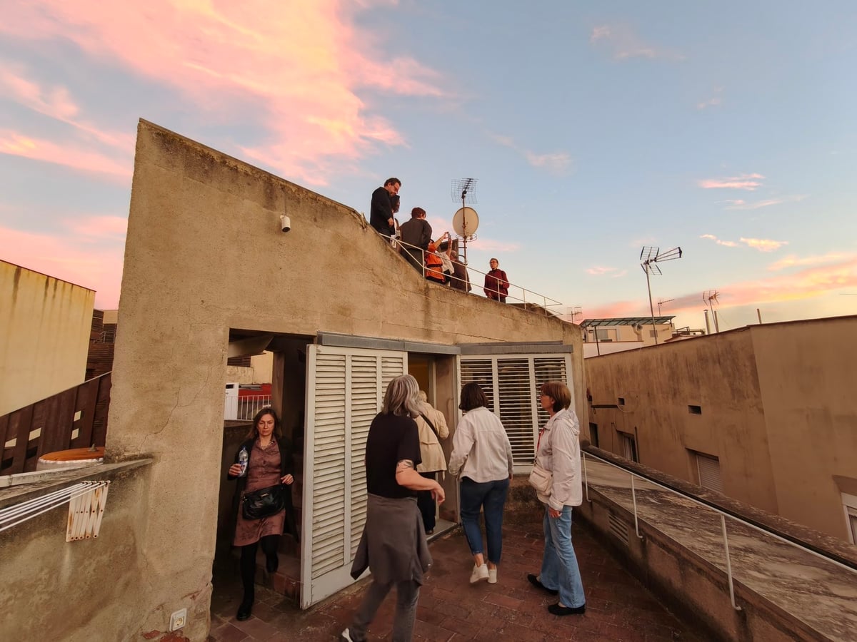 Visitantes en la terraza superior de Casa Adret, junto a una caseta de persianas de lamas y bajo el cielo rosado del atardecer.